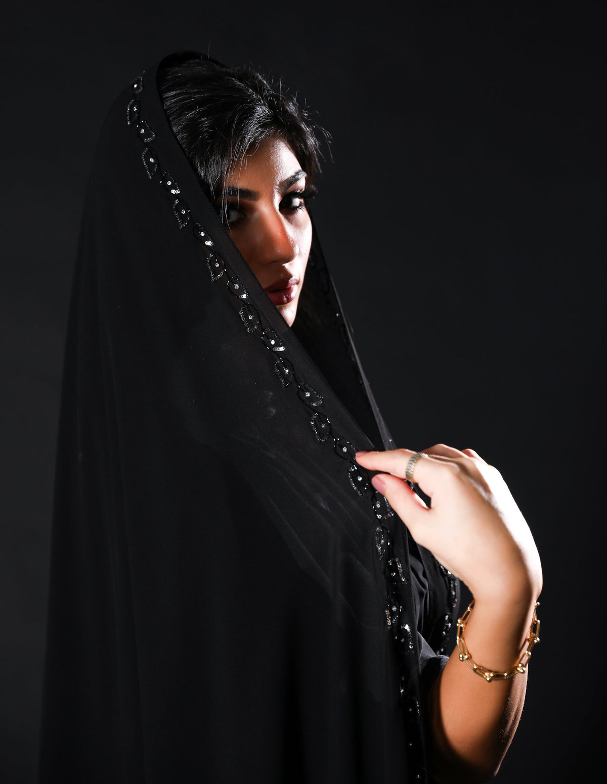 امرأة تلبس شيلة منيرة MN1-02 السوداء بشغل أسود على أطرافها أمام خلفية داكنة
